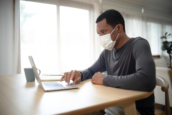 Portrait Of Handsome African American Man With Medical Mask Whil Portrait of handsome African American man with medical mask while working at desk at home during corona virus outbreak. Concept business people working at home,Remote,freelancer.COVID-19 epidemic.